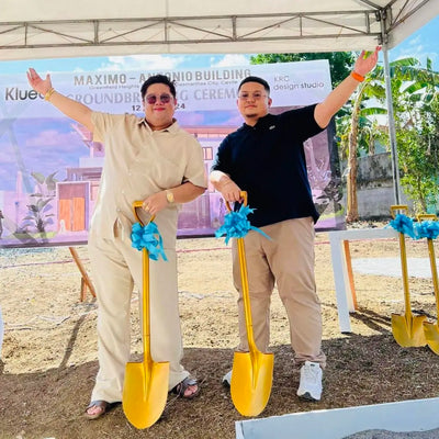 Two individuals holding yellow shovels with blue ribbons at a groundbreaking ceremony.