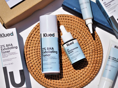 Klued skincare products including toner, serum, and moisturizer on a woven mat with packaging in the background.
